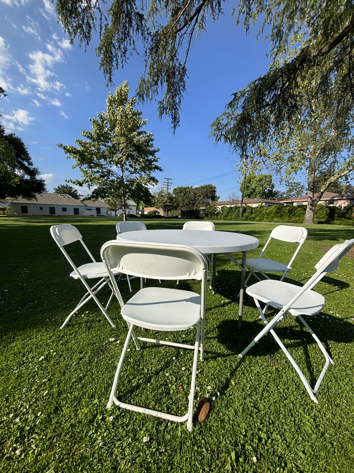Round white folding table surrounded by white folding chairs at an outdoor gathering setup